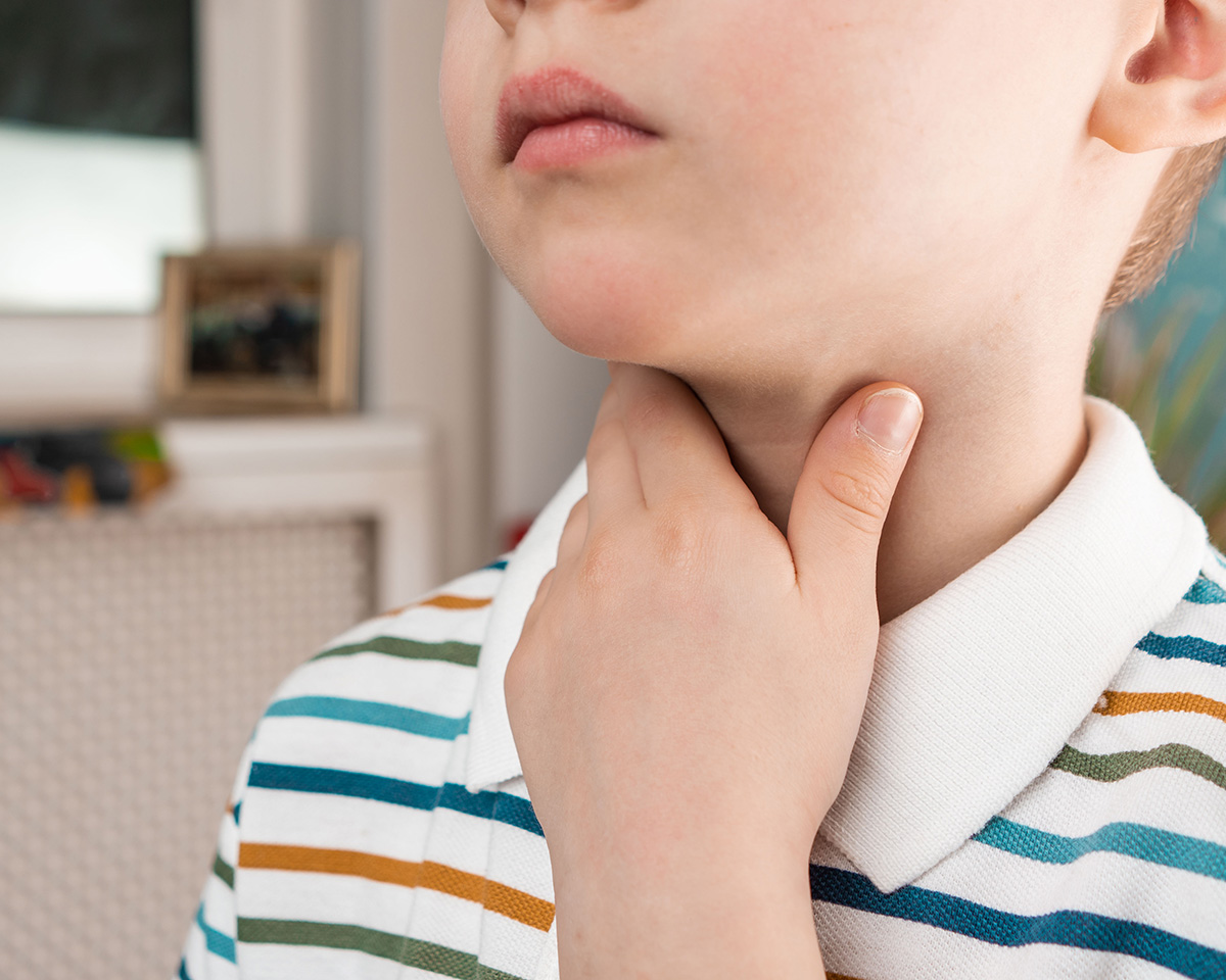 PX-RHE-LOG_Stimmstoerung-Kinder_405564541 Ein Junge mit gestreiftem Hemd berührt seinen Hals, möglicherweise wegen einer Stimmstörung.